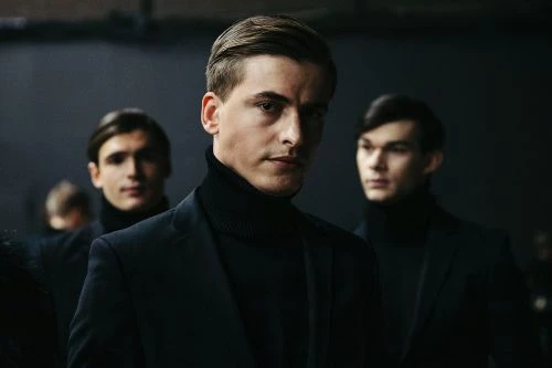 Three adult male fashion models in black turtleneck sweaters and blazers pose indoors against a dark background in a medium shot.