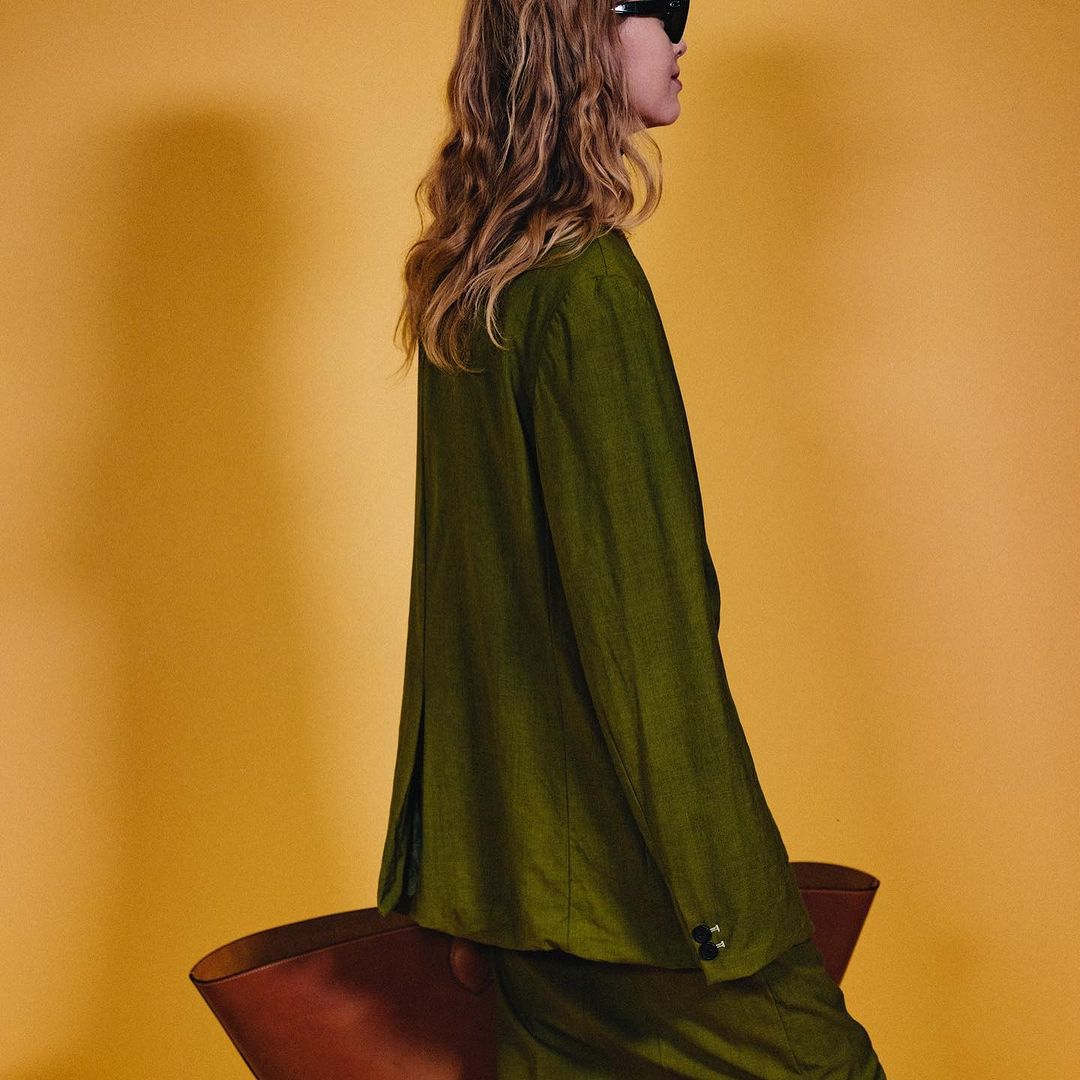 Adult female in an olive-green suit and sunglasses poses with her hand on a brown chair against a solid yellow backdrop, studio lit.