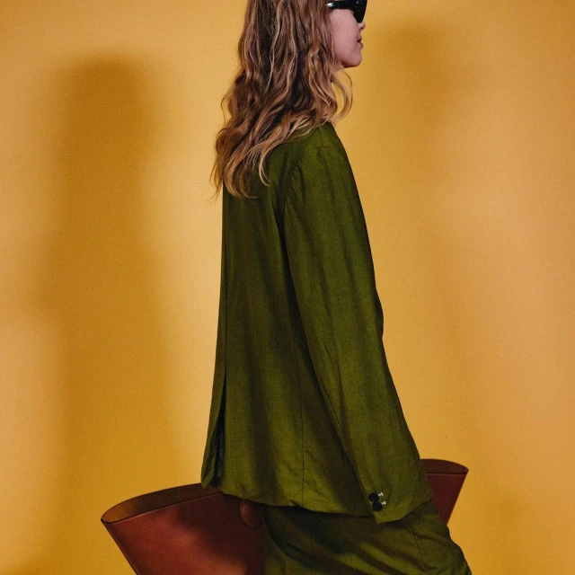 Adult female in an olive-green suit and sunglasses poses with her hand on a brown chair against a solid yellow backdrop, studio lit.