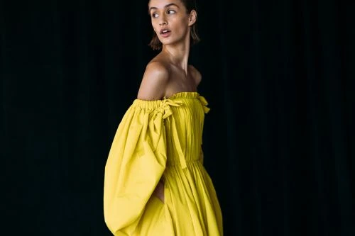 Woman in a yellow off-the-shoulder dress looking over her shoulder against a dark background