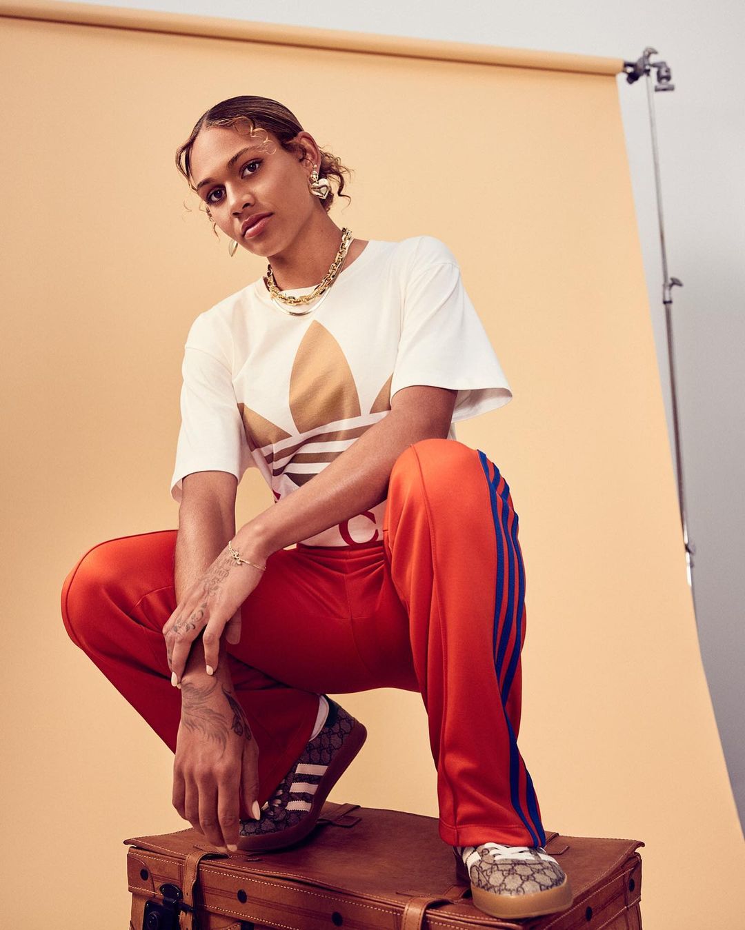 Woman in Adidas T-shirt and track pants poses in studio on brown trunk, adorned with gold jewelry, against a neutral backdrop.