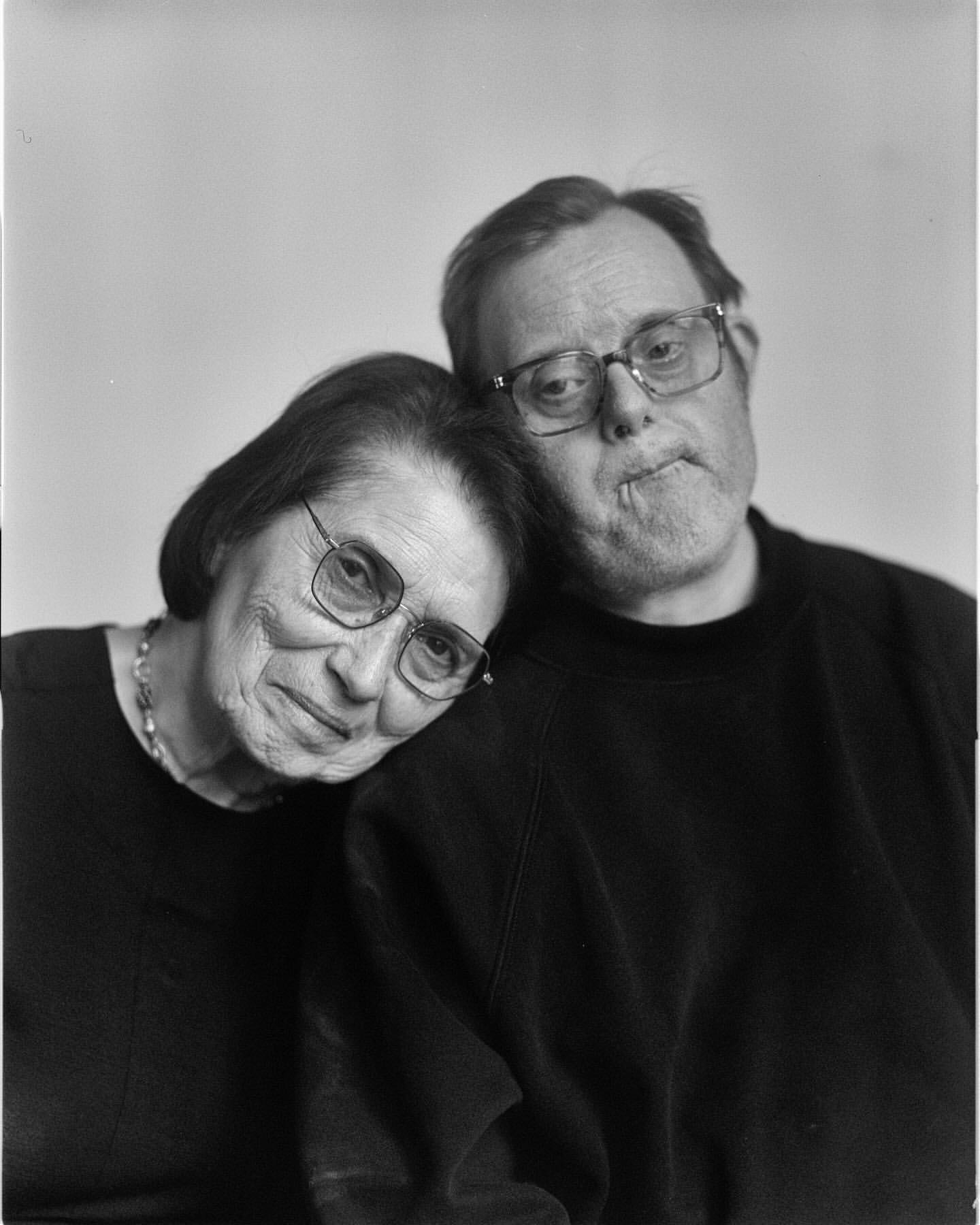 Portrait of an older woman resting her head gently on the shoulder of a man wearing eyeglasses, captured in monochrome.