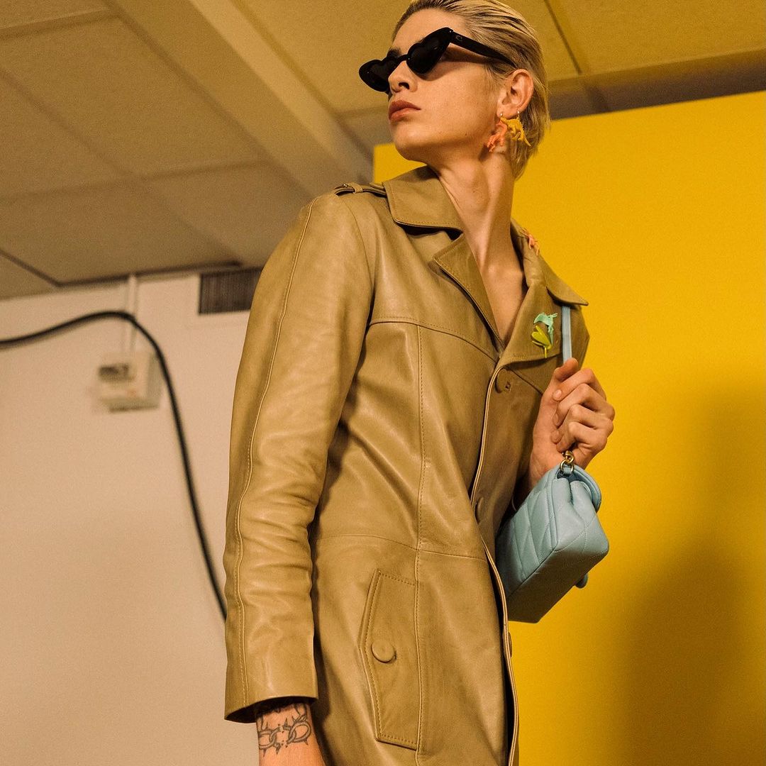A fair-skinned person in a trench coat and sunglasses poses in front of a yellow wall with a small purse and large, colorful earrings.