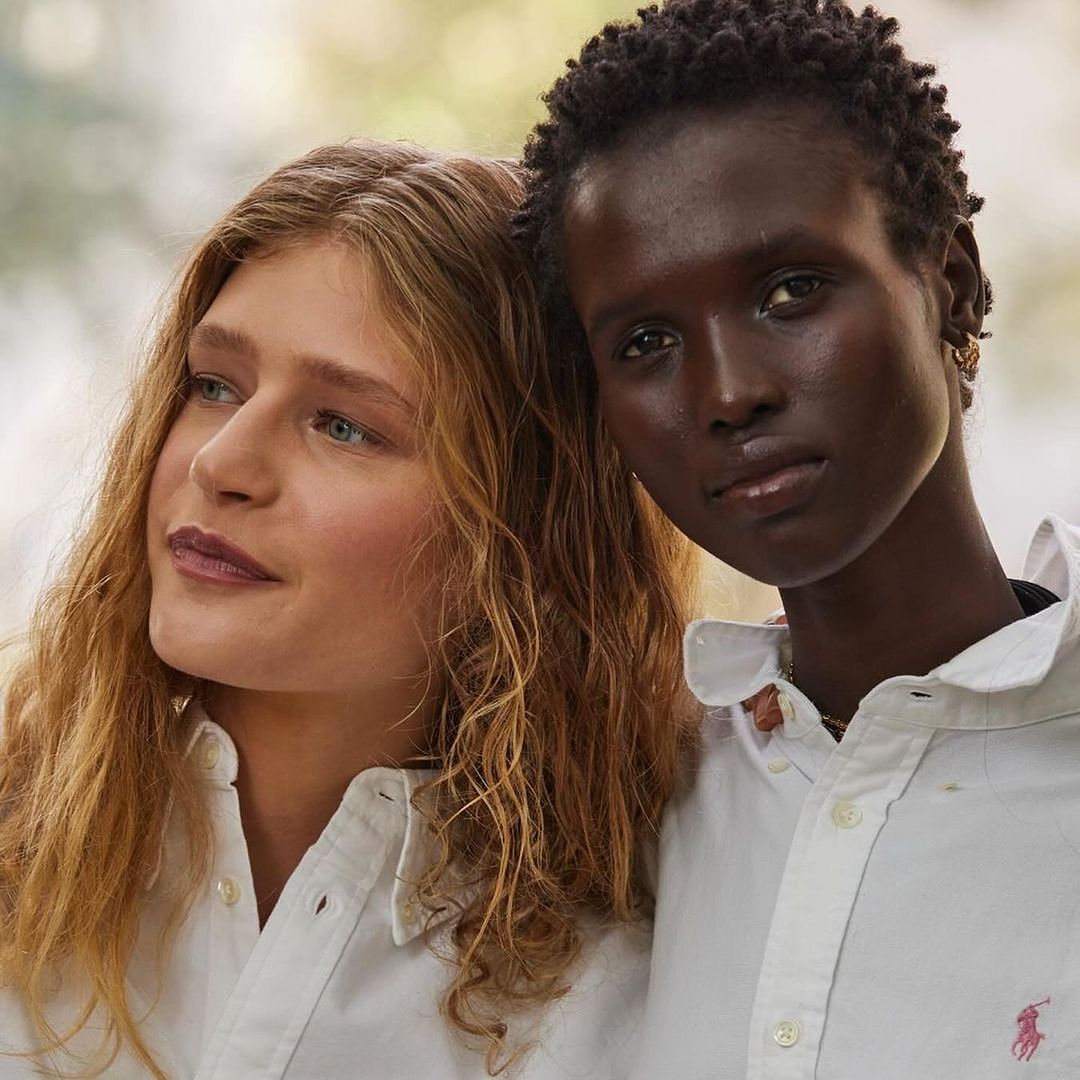 Two women of different ethnicities pose in white collared shirts against a blurred outdoor background, showcasing fashion and diversity.