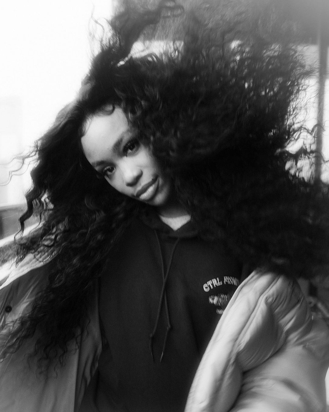 Black and white portrait of a woman with voluminous dark curly hair in motion, wearing a hooded sweatshirt and puffer jacket.
