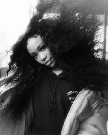 Black and white portrait of a woman with voluminous dark curly hair in motion, wearing a hooded sweatshirt and puffer jacket.