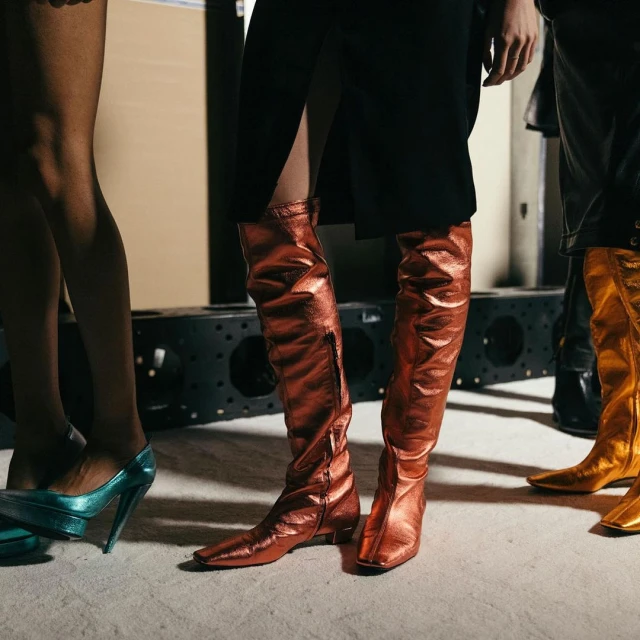 Several women model colorful, shiny thigh-high boots in a studio. The boots are metallic in teal, copper, and gold tones.