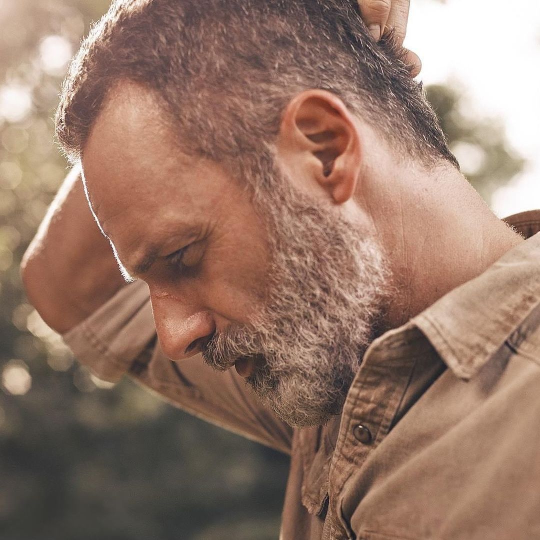 Portrait of a middle-aged adult male with a beard, wearing a button-down shirt, standing outside surrounded by foliage and sunlight.