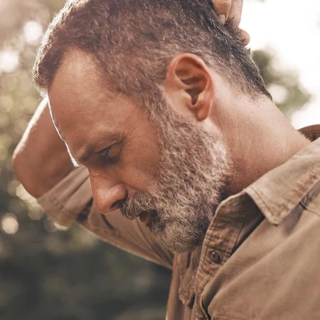 Portrait of a middle-aged adult male with a beard, wearing a button-down shirt, standing outside surrounded by foliage and sunlight.
