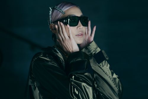 Woman in futuristic sunglasses and olive jacket poses in front of dark backdrop with hands near face in a beauty shot.