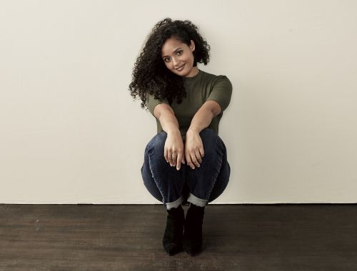 A woman with curly hair sits on the floor, wearing a green shirt, blue jeans, and black boots, posing against a cream-colored wall.