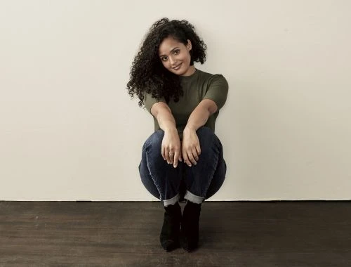 A woman with curly hair sits on the floor, wearing a green shirt, blue jeans, and black boots, posing against a cream-colored wall.
