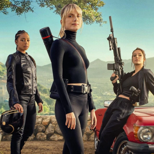 Three women stand outdoors, one in a black turtleneck, one with a motorcycle helmet, and one leaning on a vintage red convertible car.