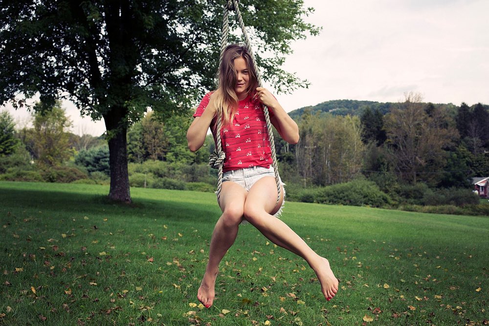 Young adult woman swings barefoot on a rope swing hanging from a tree in a grassy yard, with a backdrop of trees and a distant hill.