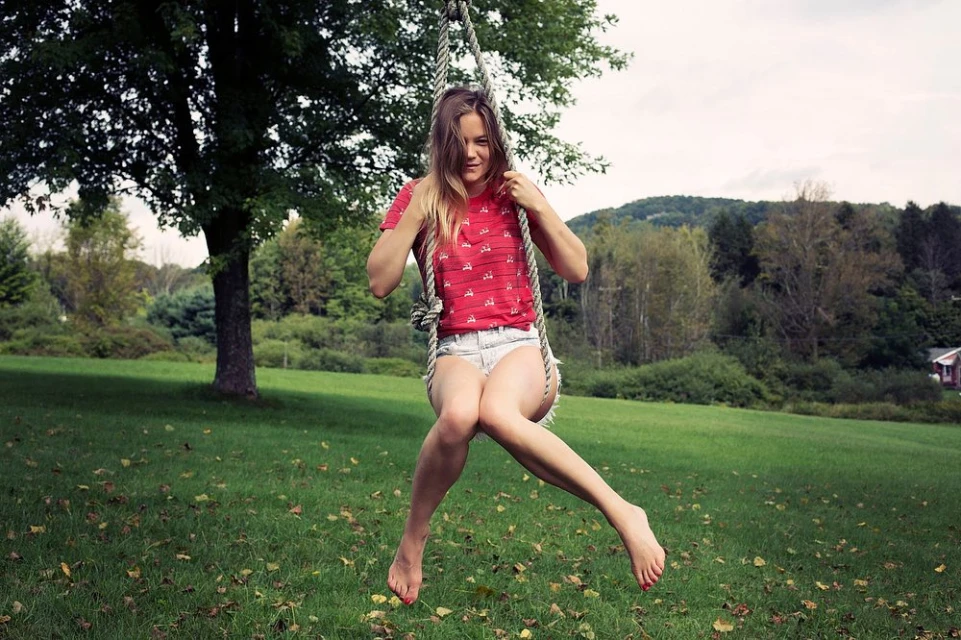 Young adult woman swings barefoot on a rope swing hanging from a tree in a grassy yard, with a backdrop of trees and a distant hill.
