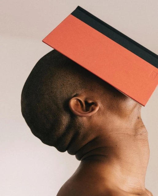 Conceptual image of a bare-chested, dark-skinned adult leaning their head to the side while balancing a coral and black book on top.