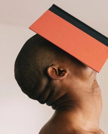 Conceptual image of a bare-chested, dark-skinned adult leaning their head to the side while balancing a coral and black book on top.
