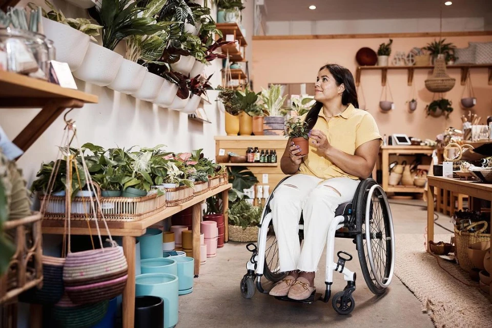 Woman in wheelchair holding potted plant at indoor plant store with shelves of plants, pots, and woven baskets. Indoor daylight.