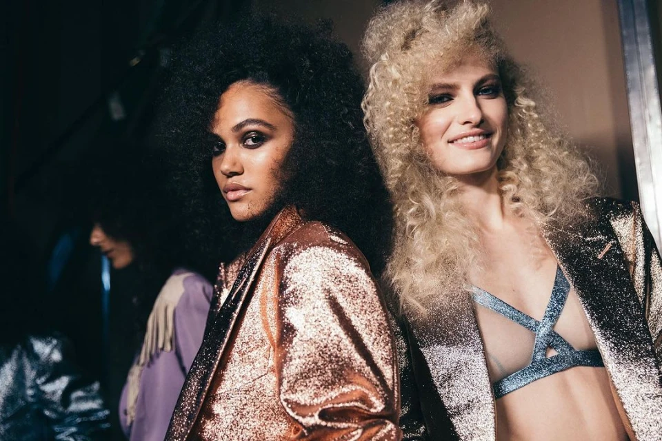 Two fashion models with curly hair in shiny jackets pose indoors, showcasing retro-inspired beauty and style for a backstage fashion moment.