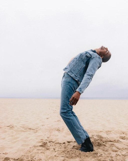 Man in denim jacket and jeans leans back on a sandy beach under an overcast sky, creating a casual and serene fashion editorial shot.