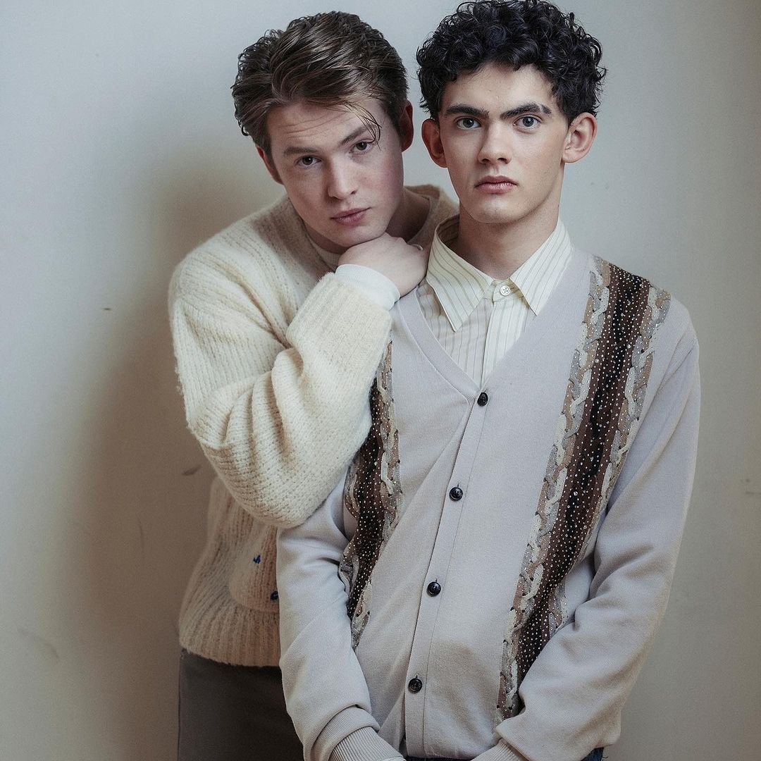 Two young men pose indoors wearing cardigans and collared shirts, one resting a hand on the shoulder of the other.