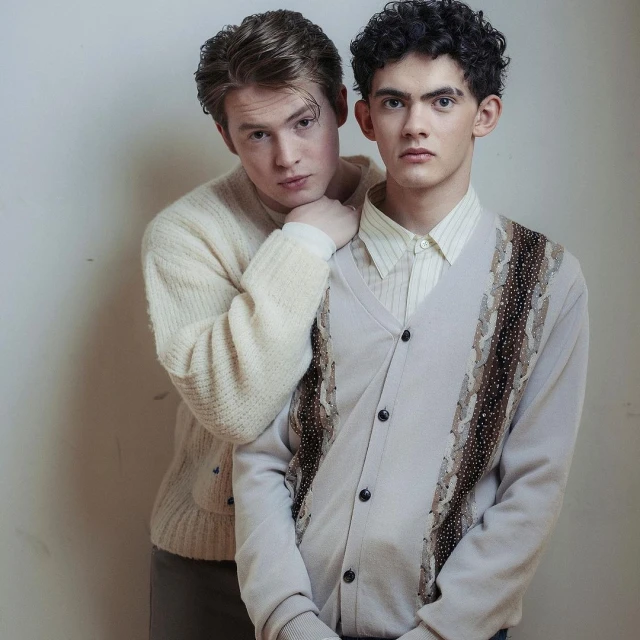 Two young men pose indoors wearing cardigans and collared shirts, one resting a hand on the shoulder of the other. 