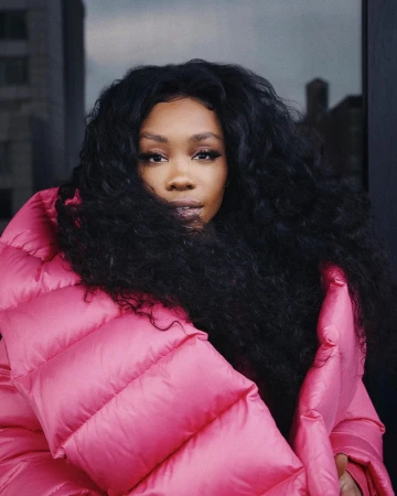 Portrait of SZA with voluminous dark hair wearing a vibrant pink puffer jacket outdoors in front of a building facade