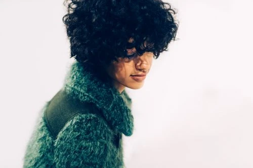 Portrait of a person with dark curly hair wearing a textured green fluffy coat against a plain white background.