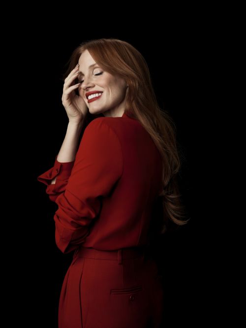 Portrait of an adult woman with red hair, wearing red attire, smiling with closed eyes, and a black backdrop.