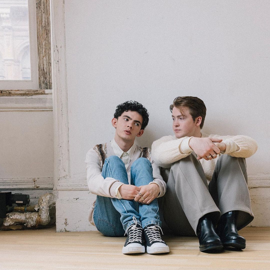 Two young men sit on a wooden floor against a white wall. One wears jeans and a white shirt, the other gray pants and a white sweater.