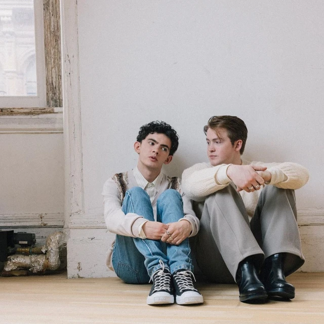 Two young men sit on a wooden floor against a white wall. One wears jeans and a white shirt, the other gray pants and a white sweater.