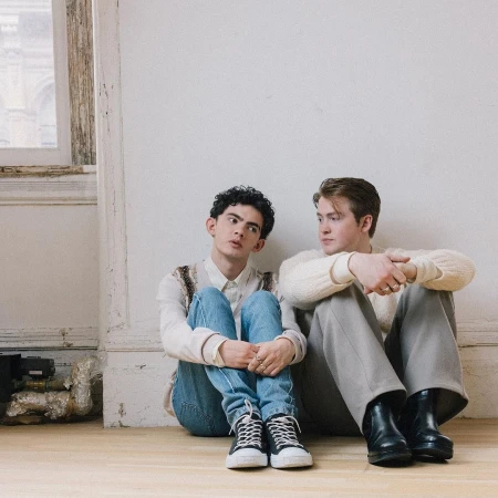 Two young men sit on a wooden floor against a white wall. One wears jeans and a white shirt, the other gray pants and a white sweater.
