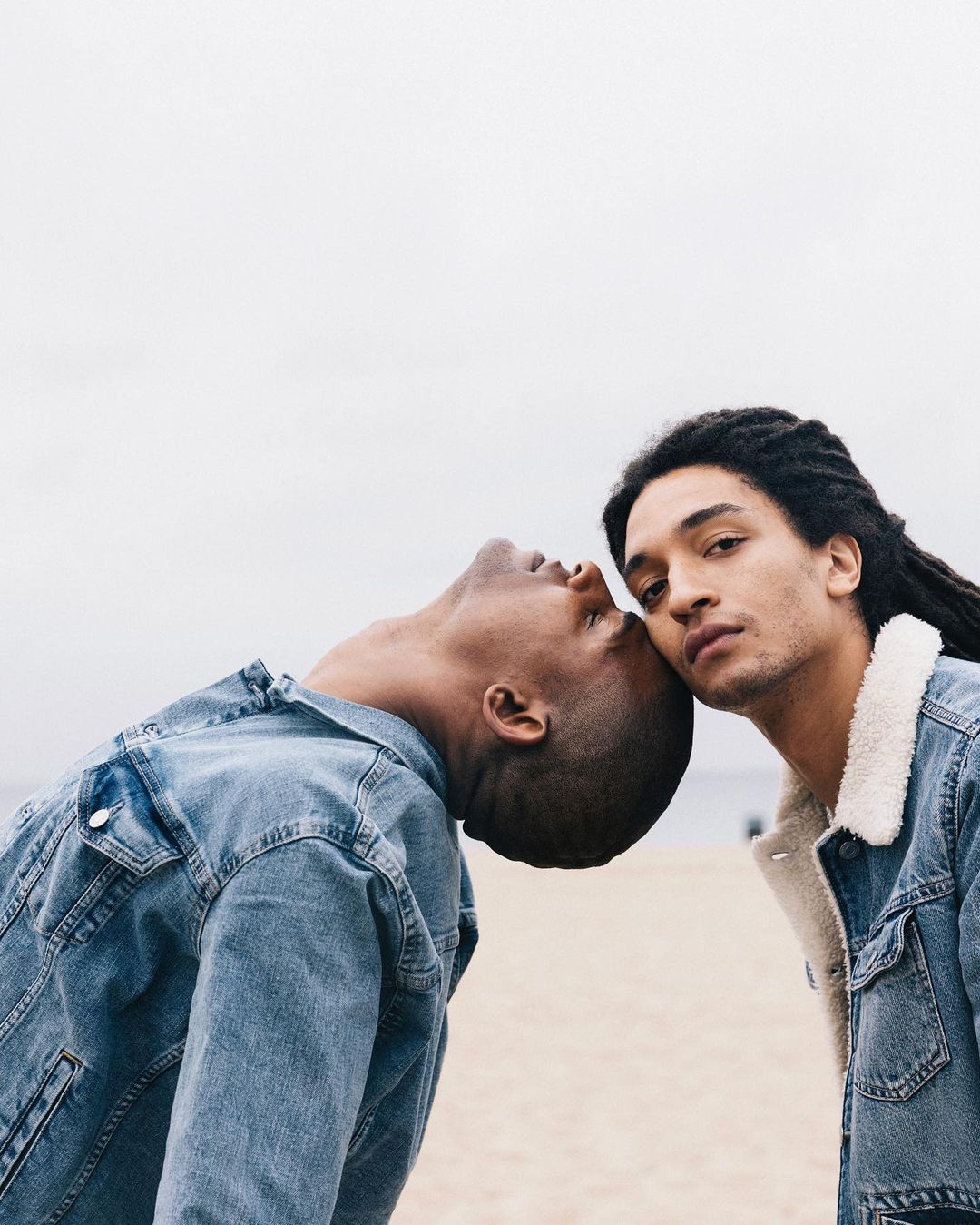 Two young men pose in denim jackets on a beach with a grey sky. One man leans his head on the other, creating a casual, intimate moment.