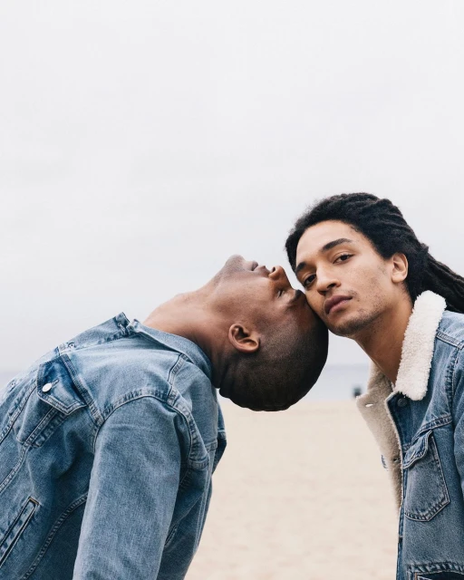 Two young men pose in denim jackets on a beach with a grey sky. One man leans his head on the other, creating a casual, intimate moment.