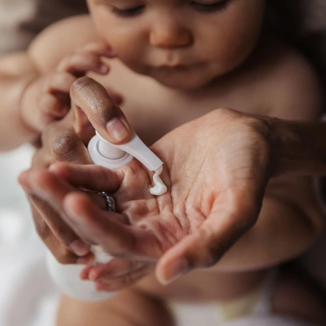Adult applying lotion to a baby's hand in an indoor setting, featuring a close-up shot with a focus on skincare and baby care.
