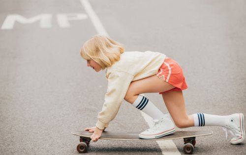 A young girl with blonde hair is skateboarding on a paved surface, wearing a yellow shirt, orange shorts, and white sneakers with knee socks.