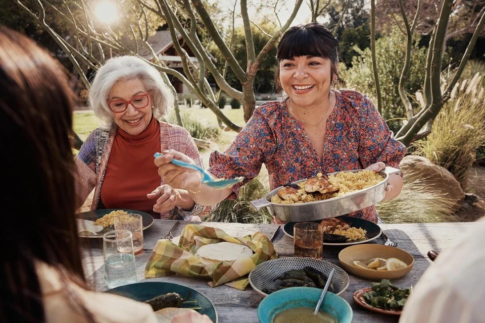 Two women enjoy an outdoor meal with family and friends. One woman holds a tray of chicken and rice, while others sit at a wooden table.