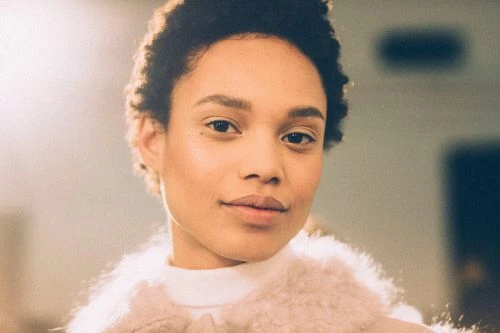 Close-up studio shot of a Black woman with short hair, smiling slightly and wearing a white turtleneck and fluffy pink outerwear.