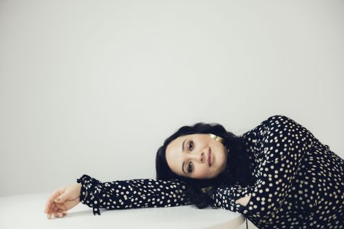 Portrait of a woman with dark hair in a black and white polka dot dress lying on a white surface, against a plain white background.