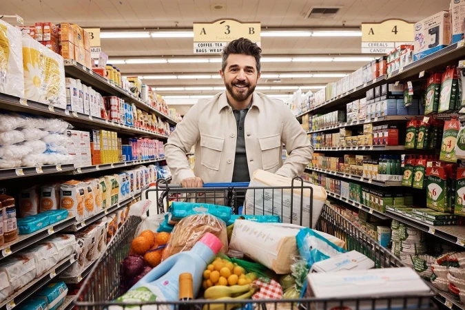 Adult man with a smile stands in a supermarket aisle, pushing a shopping cart full of groceries toward the camera in a medium shot.