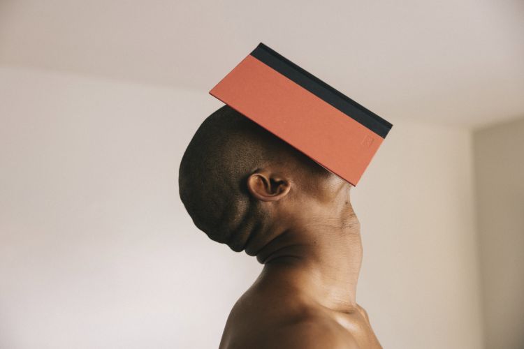 Striking studio shot of an African American adult male balancing a coral-colored book on his head against a neutral wall, close-up view.
