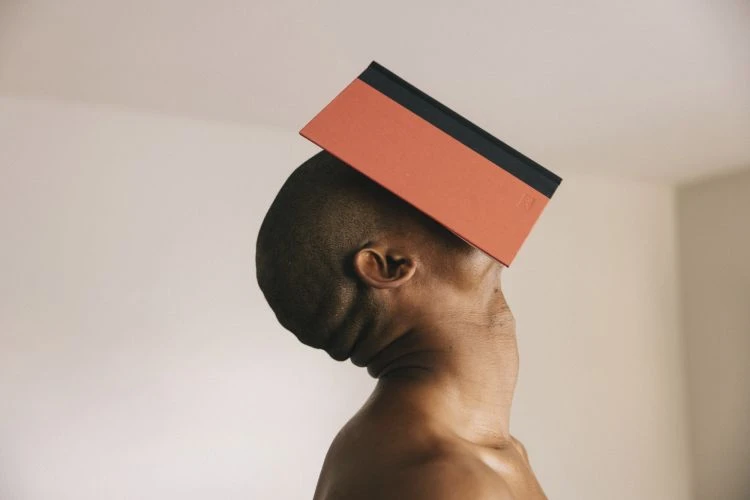 Striking studio shot of an African American adult male balancing a coral-colored book on his head against a neutral wall, close-up view.