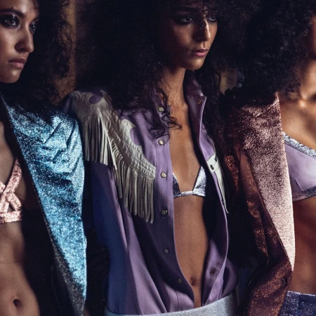 Three models with dark curly hair wearing metallic jackets and fringe shirts in a studio portrait by Victoria Will