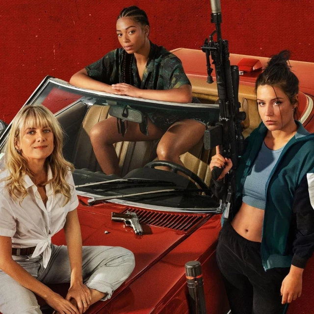 Three women pose with a red car and weaponry against a red background. One woman sits on the car hood, another inside, and another beside the car.