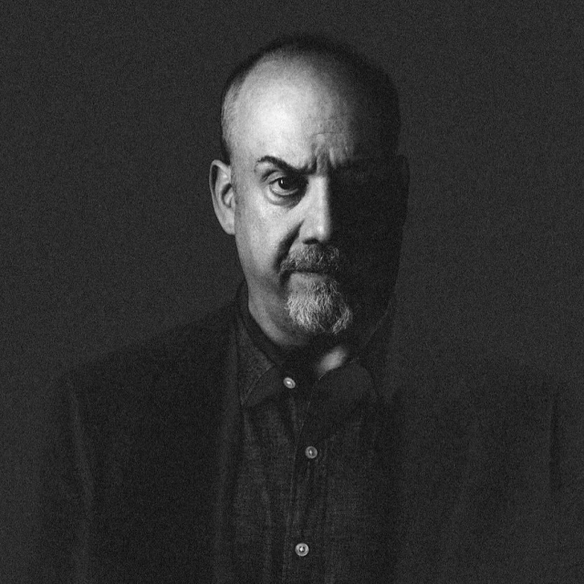 Monochrome portrait of a mature adult man with a beard wearing a collared shirt and jacket in a dark, indoor environment.