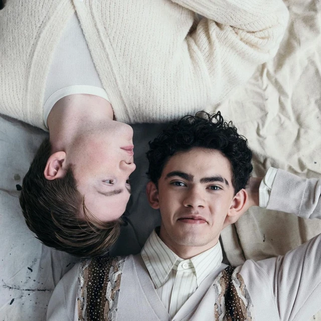 Two young men lie head-to-head, smiling, wearing light-colored sweaters against a light backdrop, portraying a relaxed, intimate moment.