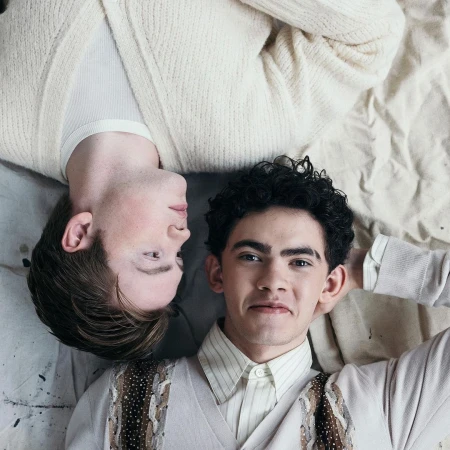 Two young men lie head-to-head, smiling, wearing light-colored sweaters against a light backdrop, portraying a relaxed, intimate moment.