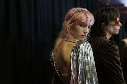 Two fashionably dressed people pose against a dark background; one with pink hair, tiara, and a silver dress, the other in a blazer.