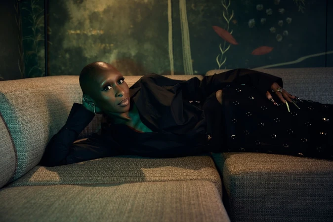 Elegant woman with shaved head poses on a couch, wearing a black outfit in a moody interior with patterned wallpaper and soft lighting.