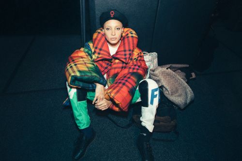 A woman with a shaved head, wearing a beret and plaid puffer jacket, is sitting on the floor with handbags in a dimly lit room.